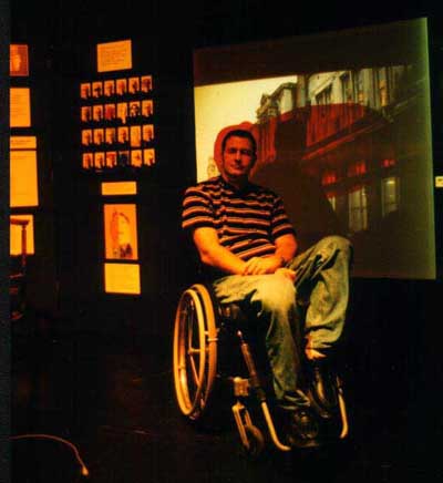 This photo was taken at a Stalking Histories workshop. A wheelchair user is on the set in front of the projection screen showing a  grey building with a large red stain over the middle section. This red stain is also across the face of the workshop participant. Photo Felicity Shillingford. 